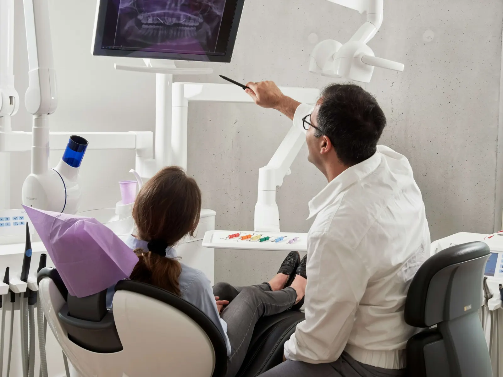 A dentist pointing to a dental X-ray on a monitor while a patient looks on